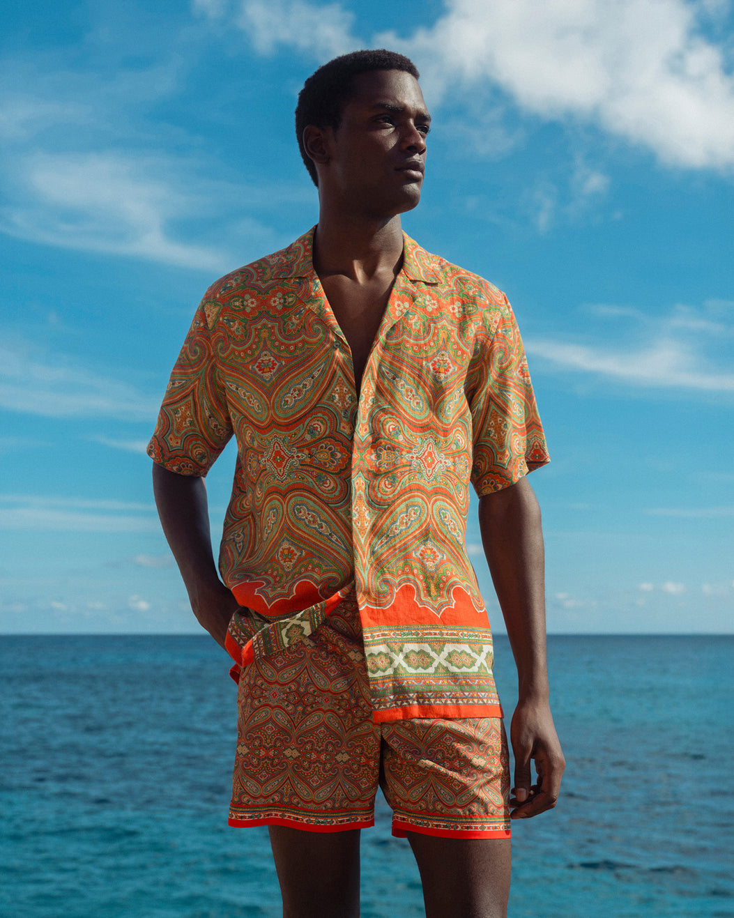 Man wearing a patterned shirt and shorts standing by the ocean with a blue sky.