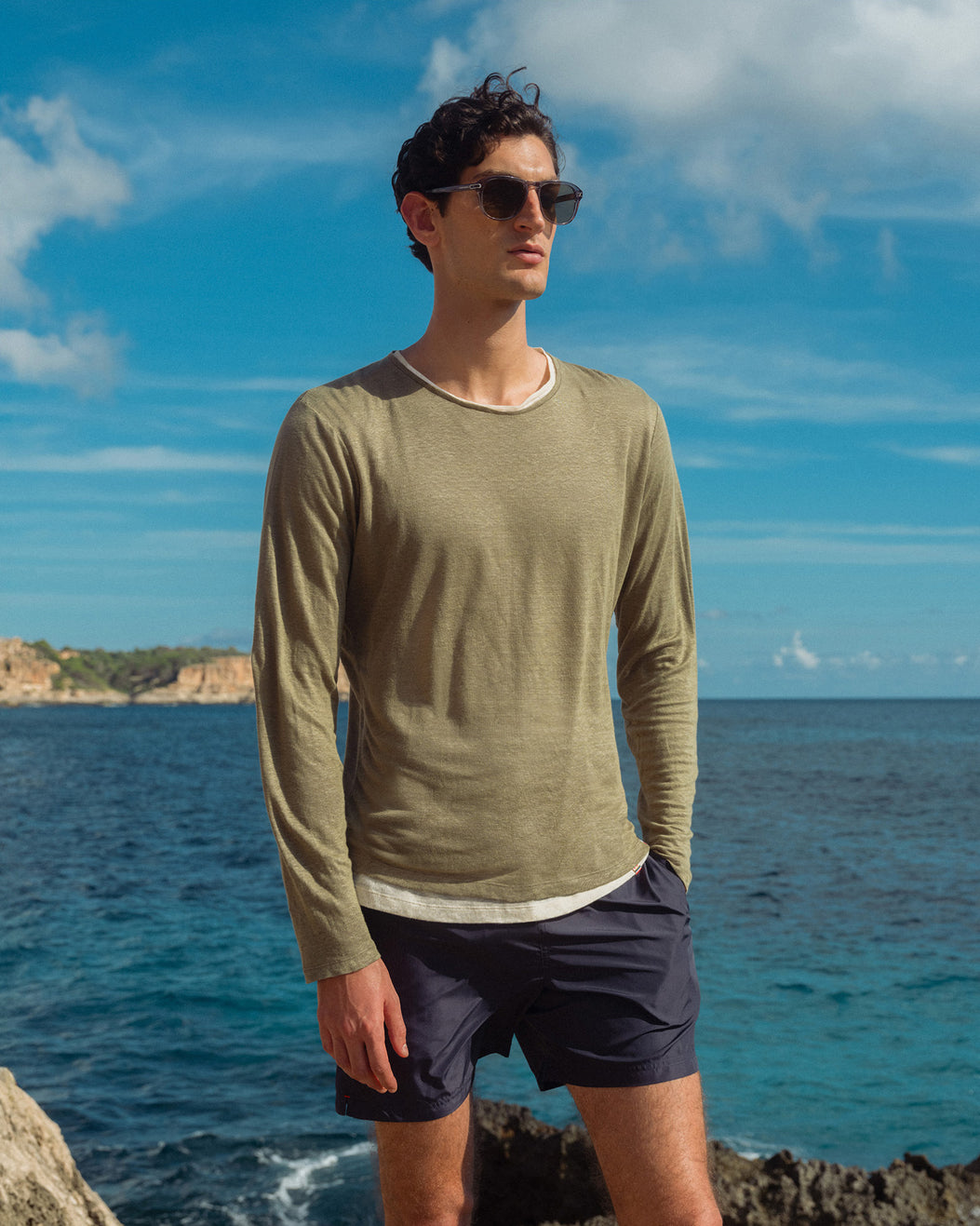 Man wearing sunglasses and a green long-sleeve shirt standing by the ocean.