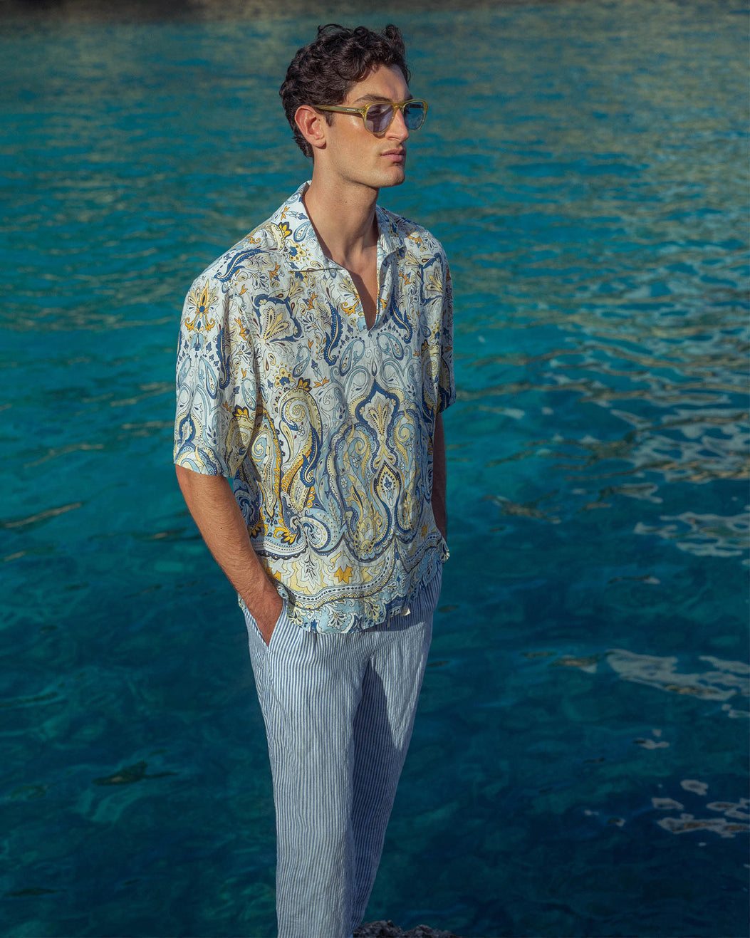 Man wearing a patterned shirt and sunglasses standing by a body of water.
