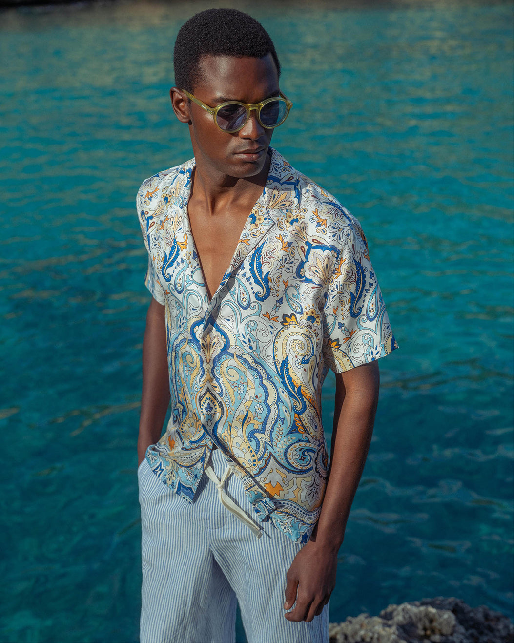 Man wearing a patterned shirt and sunglasses standing in front of blue water.