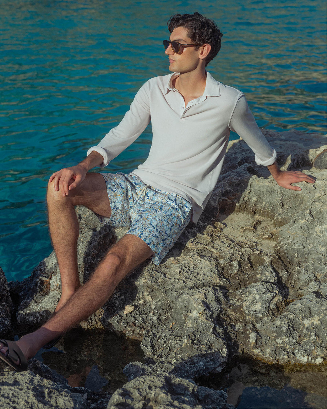 Man sitting on rocks by the water wearing a light-colored shirt and patterned shorts.