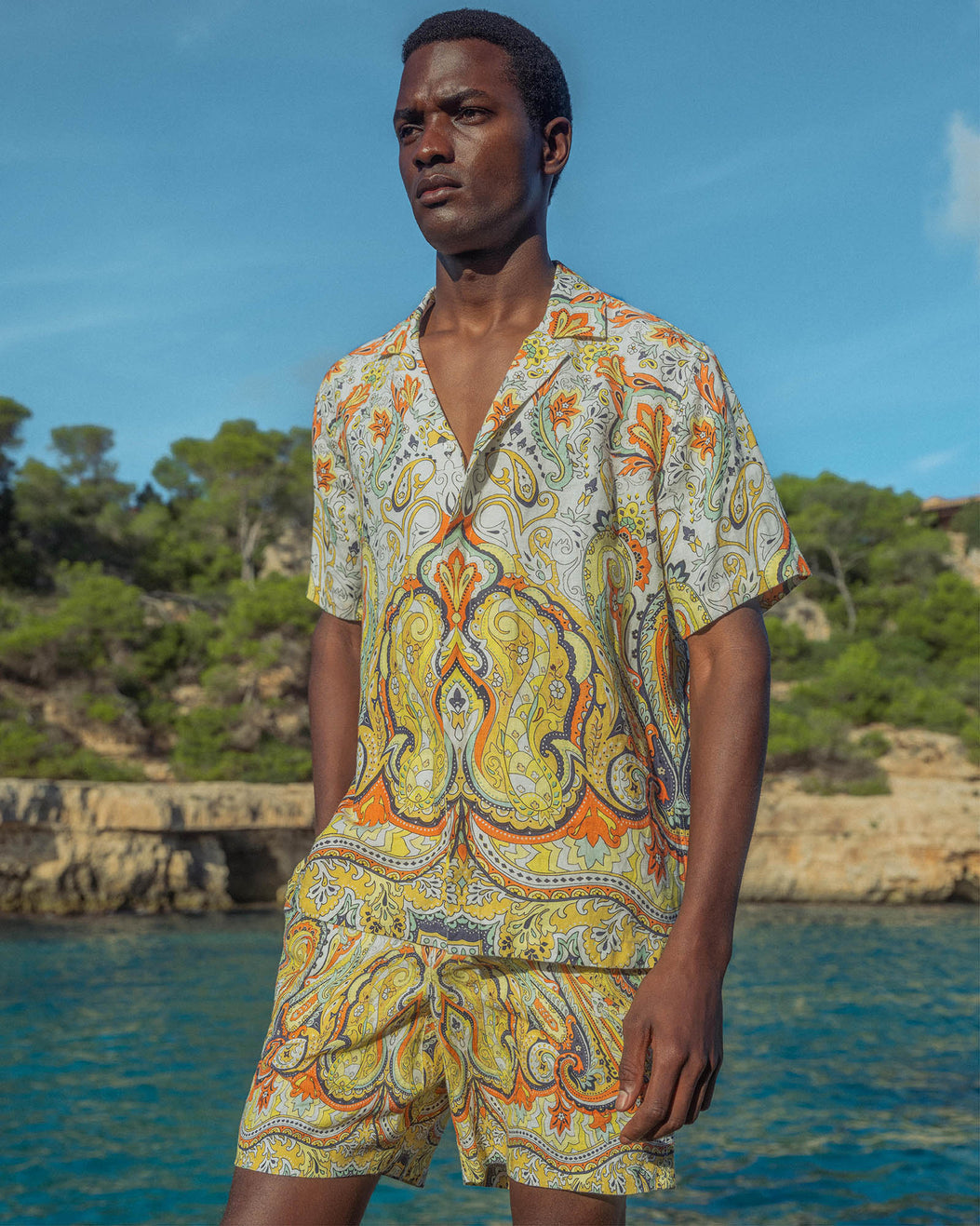 Man wearing a colorful paisley shirt and shorts standing by a body of water with greenery in the background.