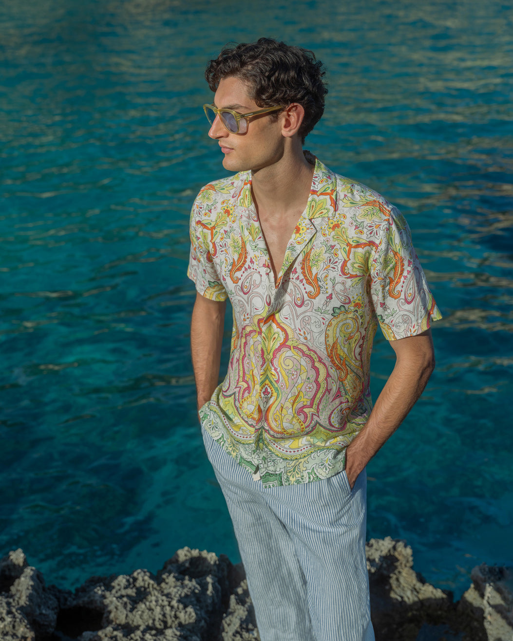 Man wearing a colorful patterned shirt and sunglasses standing by the ocean.
