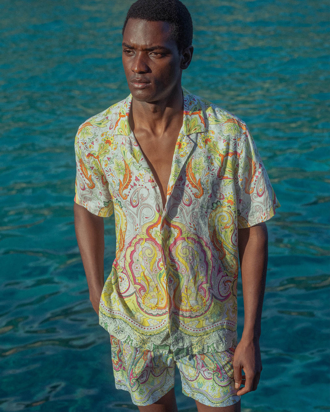 Man wearing a colorful paisley shirt and shorts standing in front of blue water.