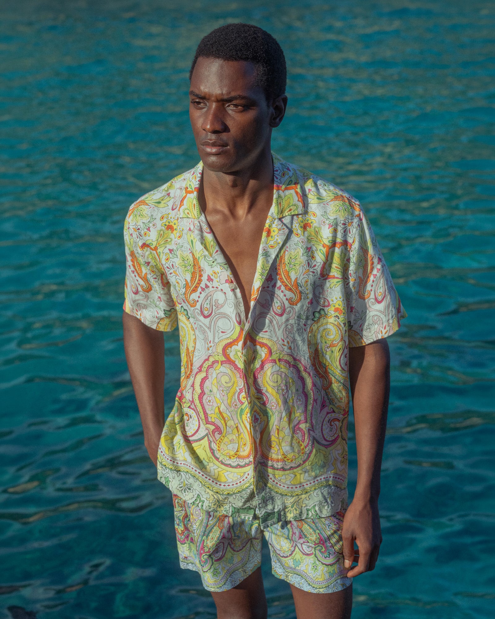 Man wearing a colorful paisley shirt and shorts standing in front of blue water.