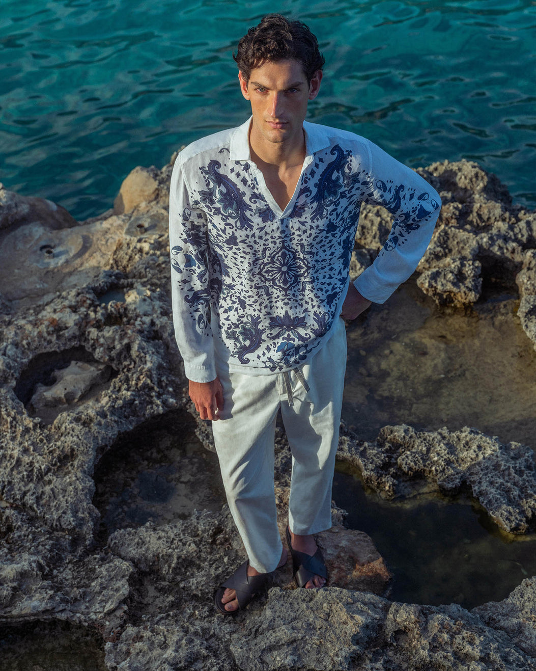 Man wearing a patterned shirt and light pants standing on rocky terrain with water in the background