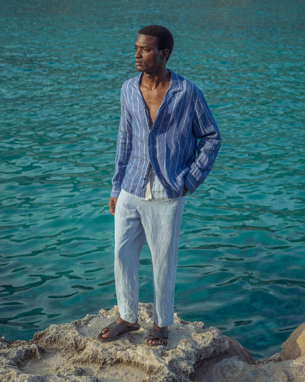 Man in a blue striped shirt and light pants standing by the ocean.