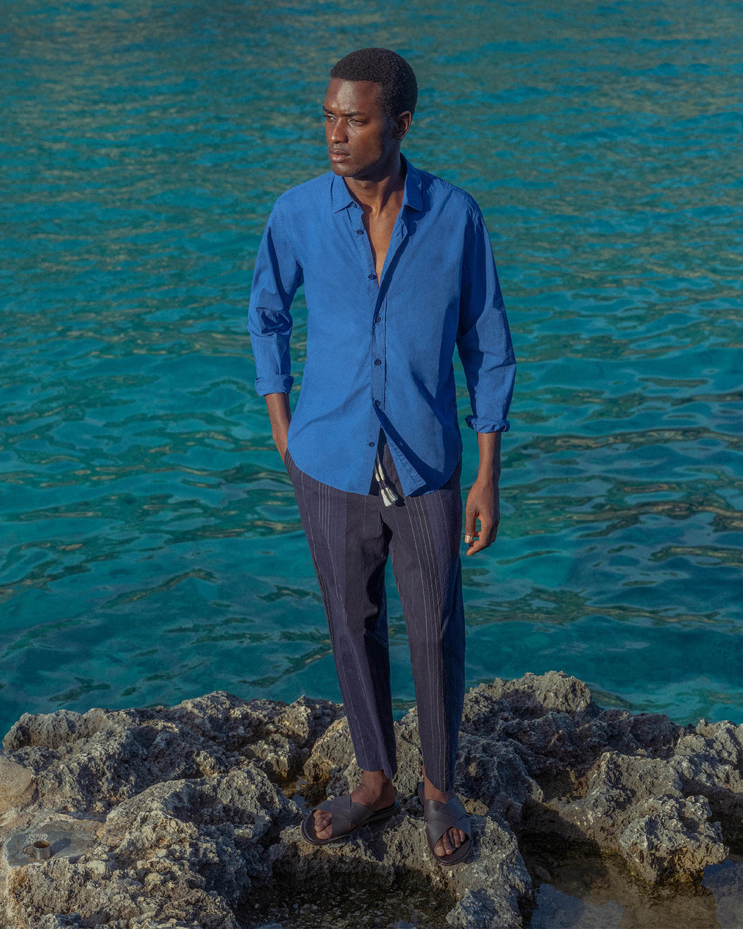 Man in a blue shirt and dark pants standing on rocks with turquoise water in the background