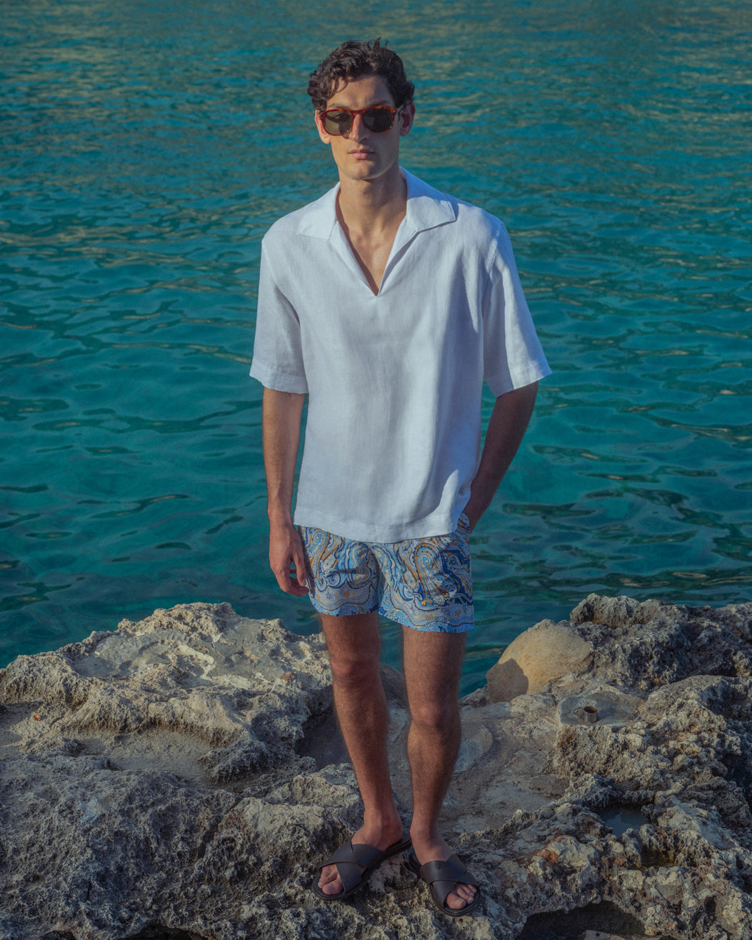 Man standing on rocky terrain by a body of water wearing sunglasses and a white shirt.