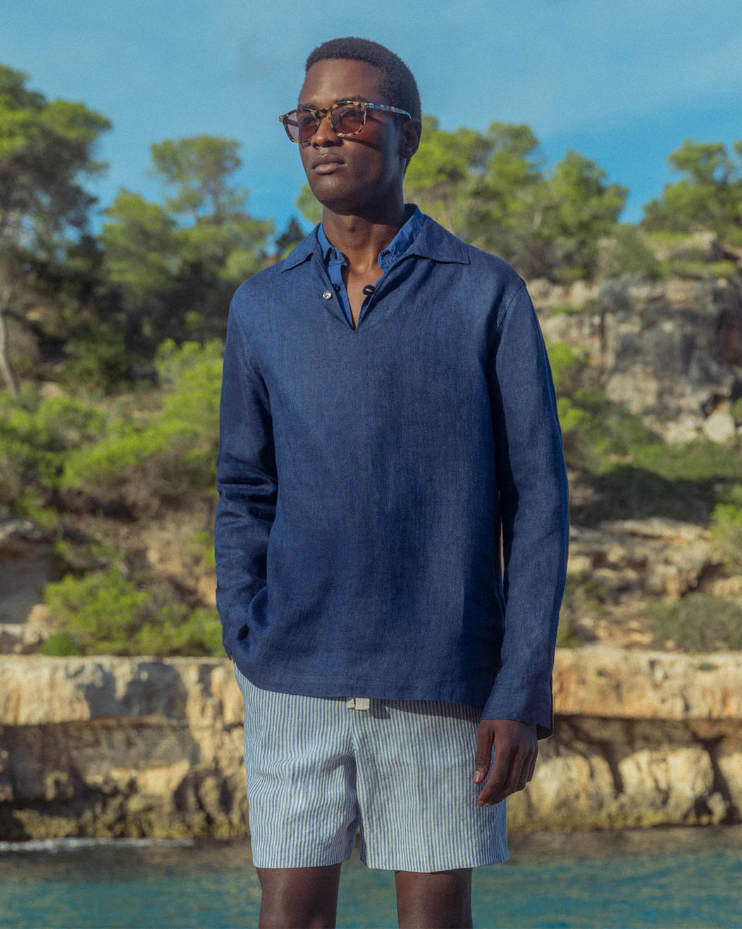 Man wearing a blue sweater and sunglasses standing by a pool with trees in the background
