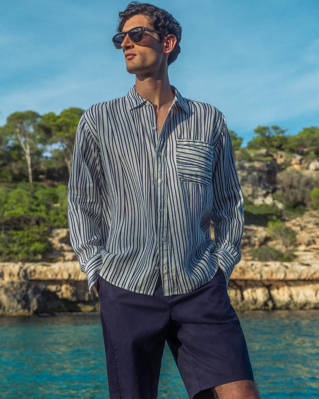 Man wearing a striped shirt and sunglasses by a body of water with greenery.