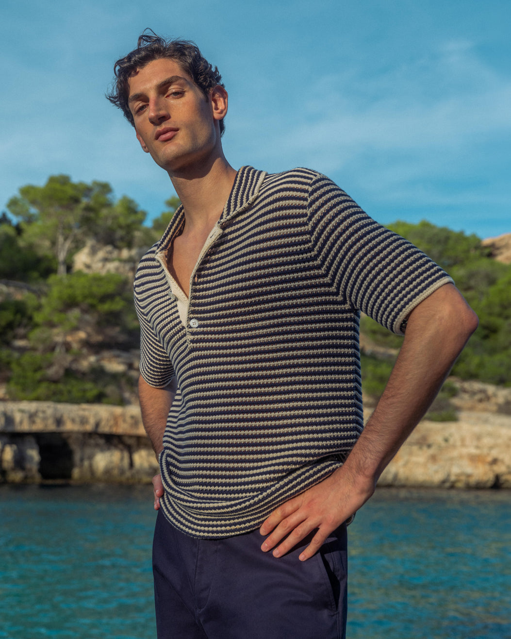Man wearing a striped shirt and dark shorts standing by a body of water with trees in the background