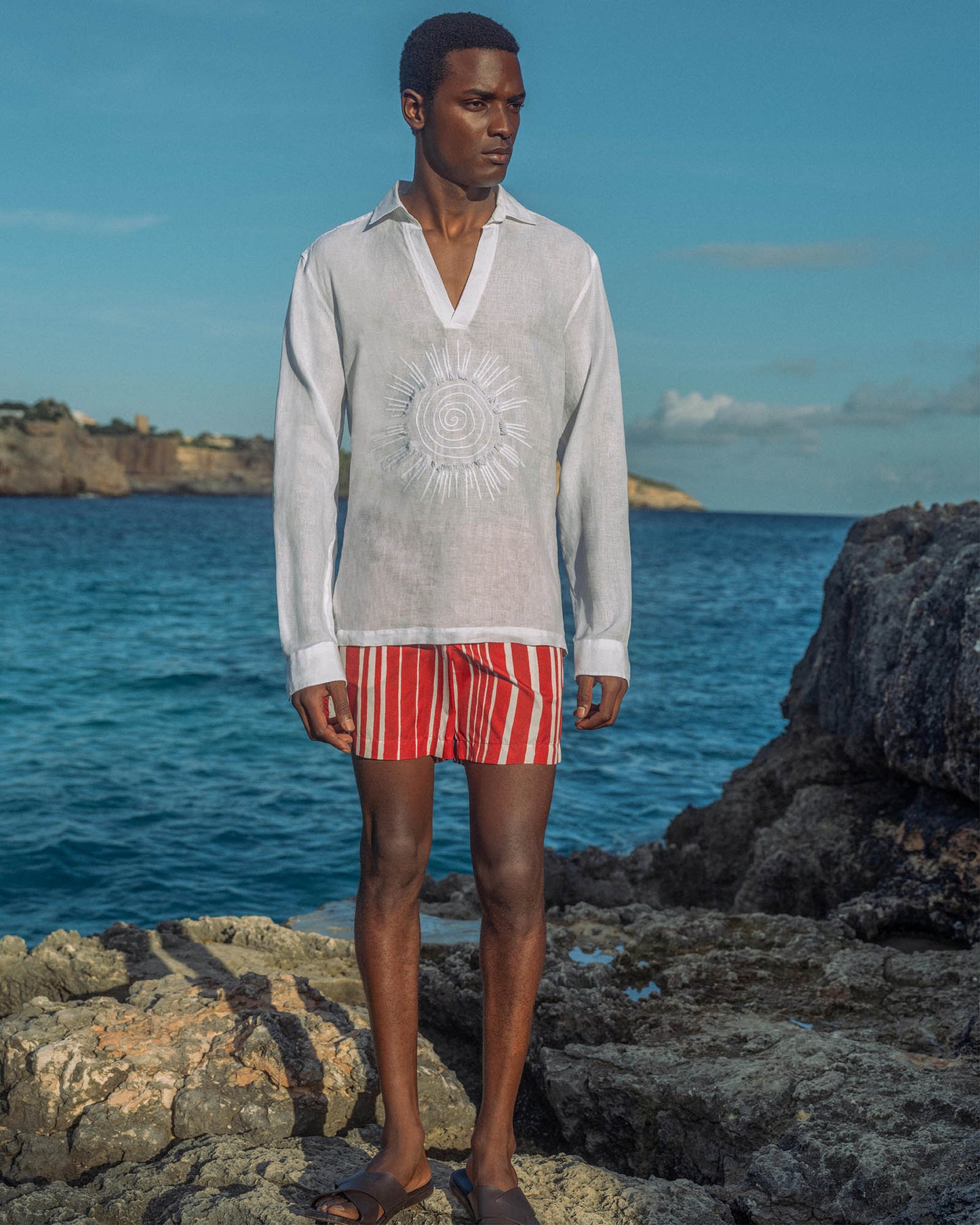 Man standing on rocks by the ocean wearing a light shirt and red striped shorts.