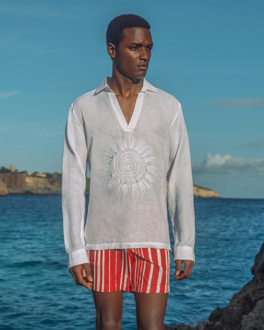 Man wearing white shirt with sunshine embroidery and red swim shorts standing by the ocean