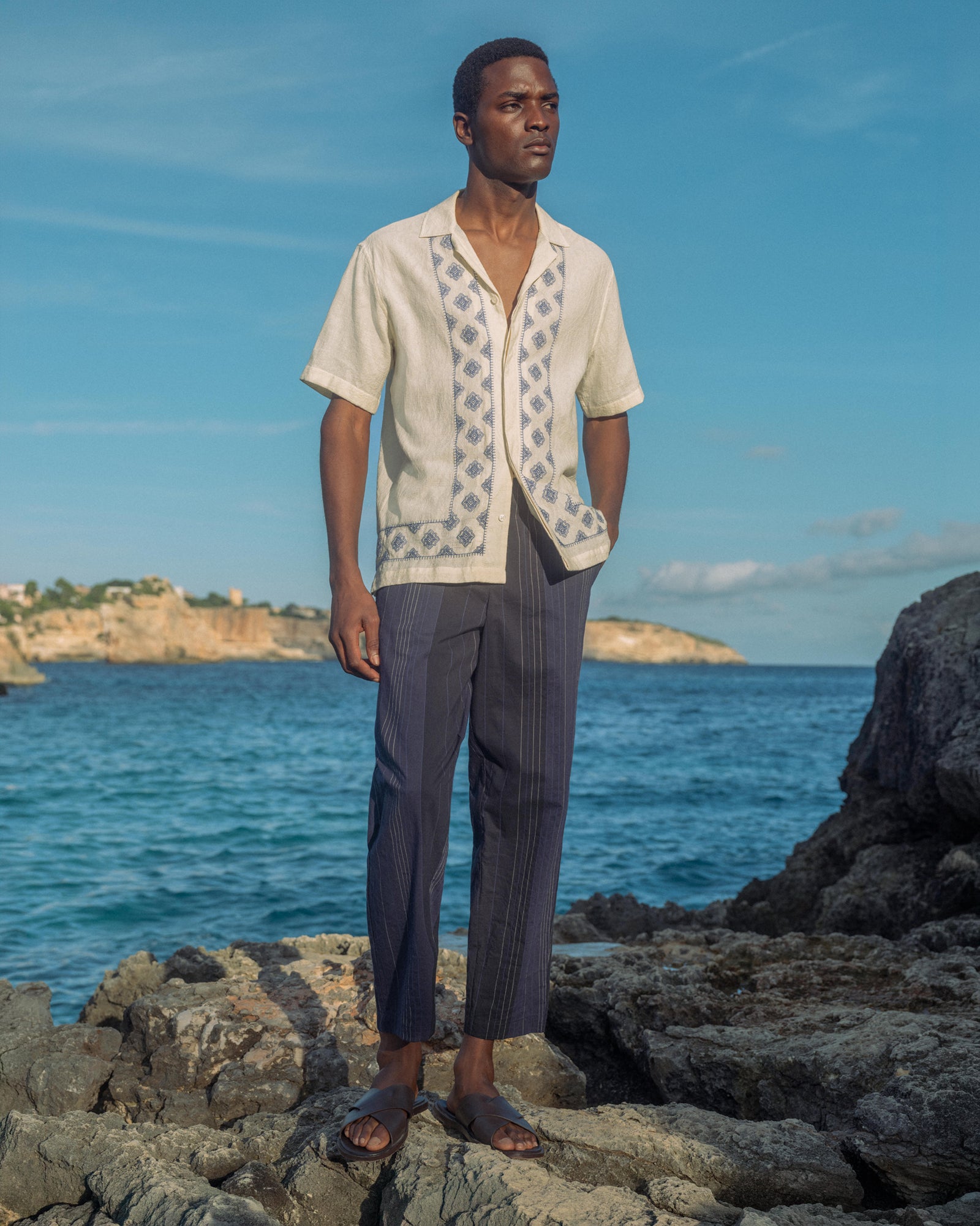 Man standing on rocks by the ocean wearing a light-colored shirt and dark pants.