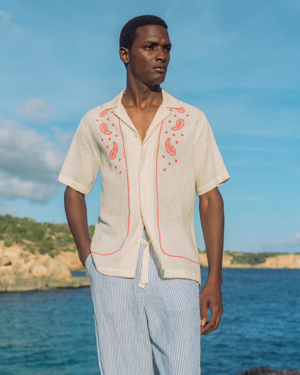 Man wearing a beige embroidered shirt with red patterns by the ocean