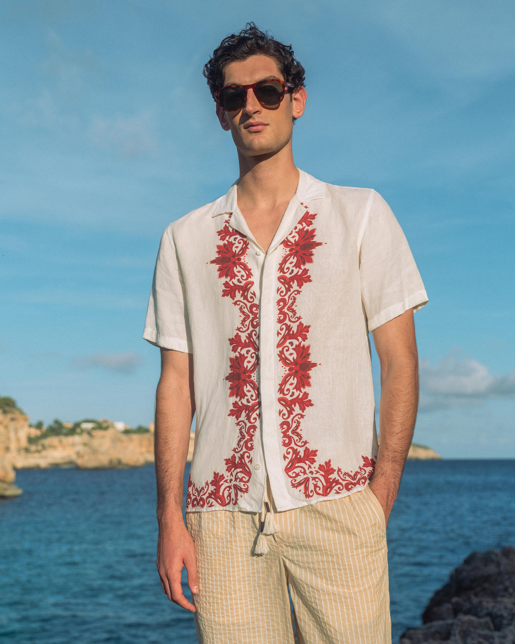 Man wearing sunglasses with white shirt with red embroidery and beige trousers standing by the ocean
