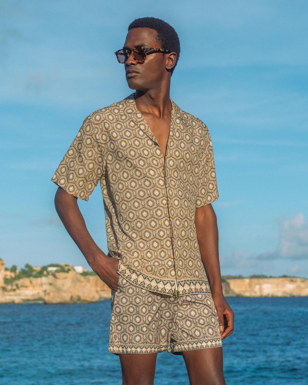 Man wearing printed shirt and shorts with sunglasses standing by the ocean