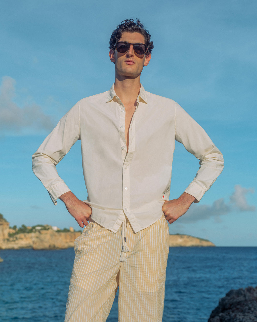 Man wearing sunglasses and a light-colored shirt and pants standing by the ocean.