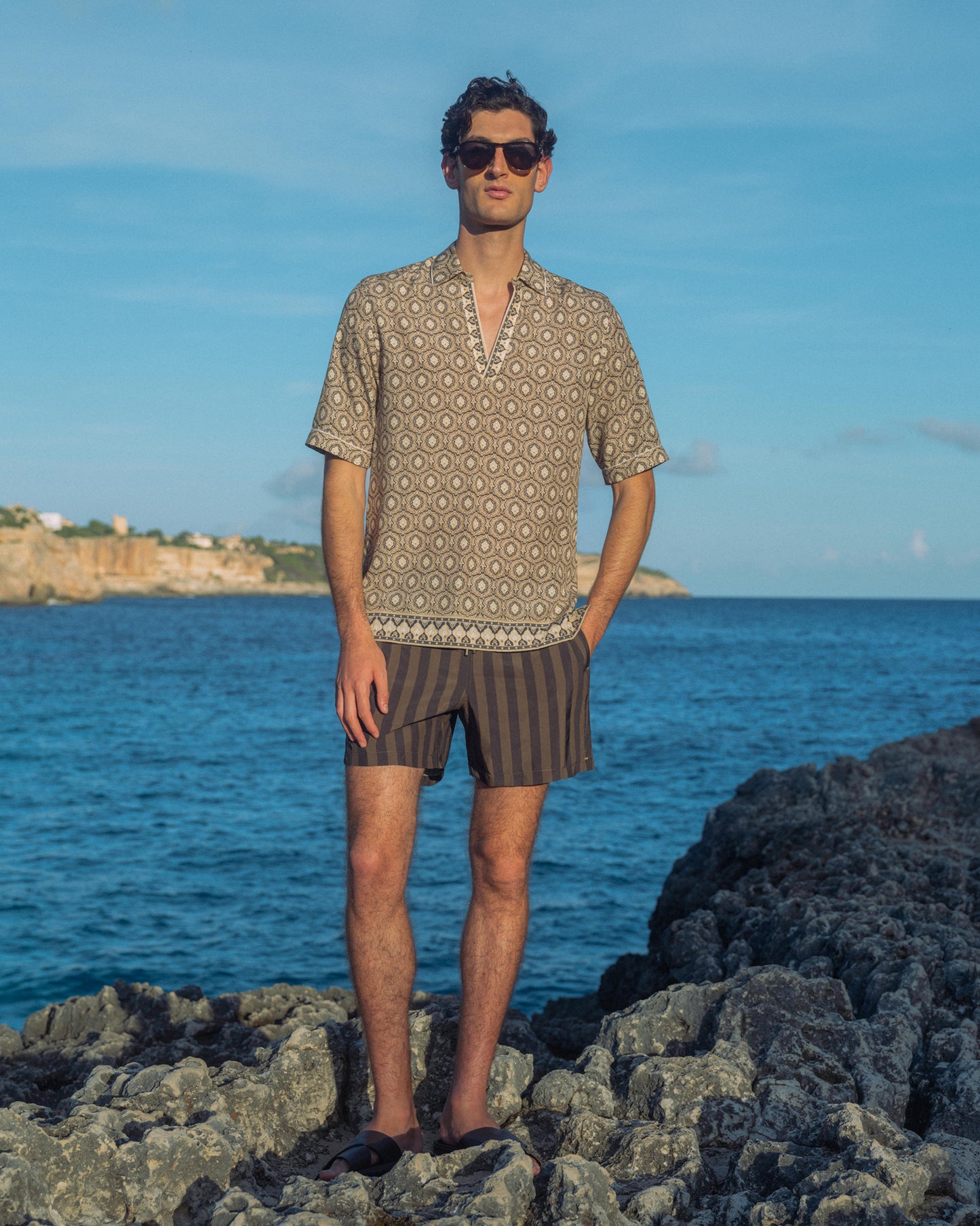 Man standing on rocks by the ocean wearing a patterned shirt and striped shorts.