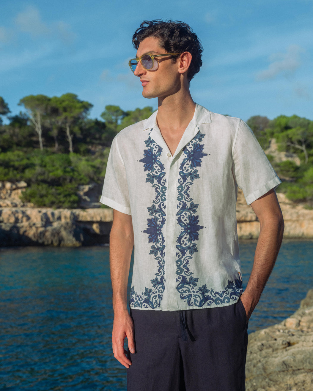 Man wearing a white shirt with blue floral embroidery by a body of water.