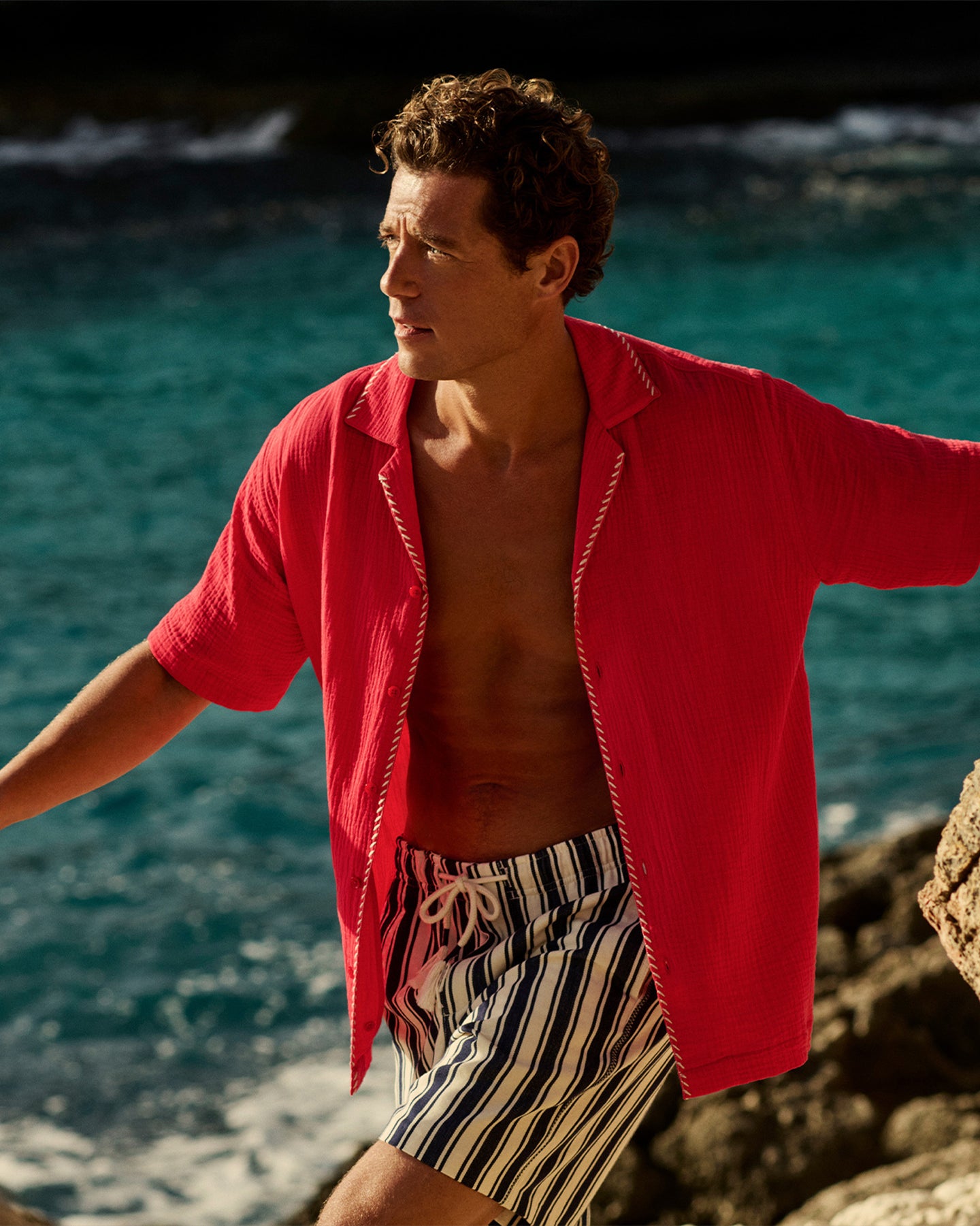 Man wearing red short sleeve shirt with striped shorts against the ocean