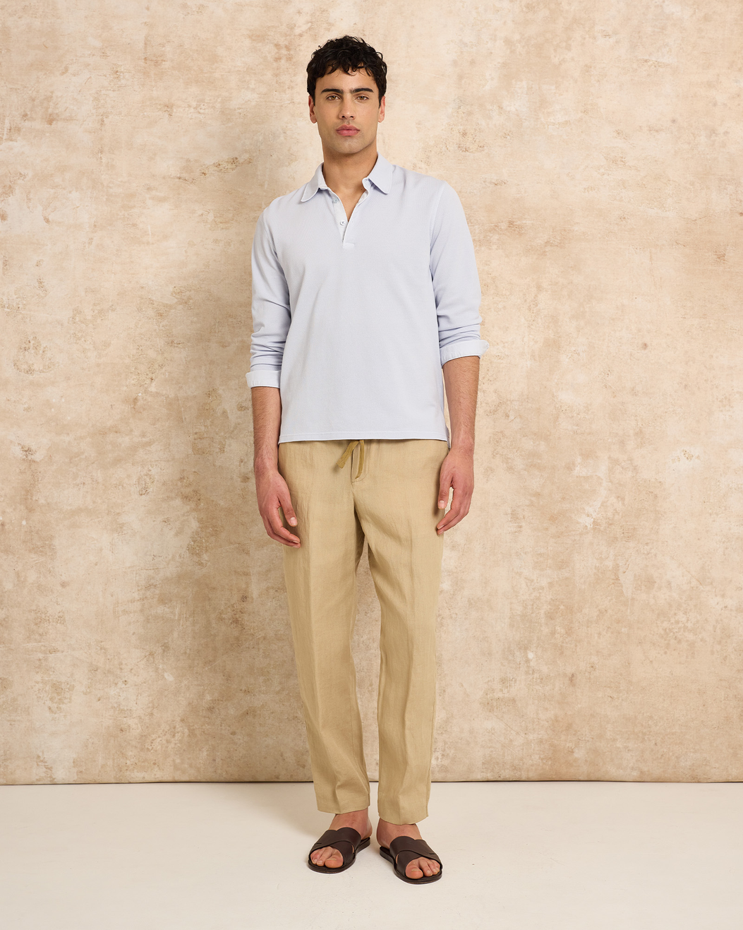 Man wearing blue long sleeve polo with beige trousers on a plain background