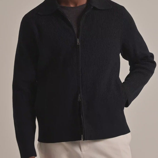 Man wearing black knitted zip-through jacket with dark grey t-shirt underneath and light beige trousers on a plain background