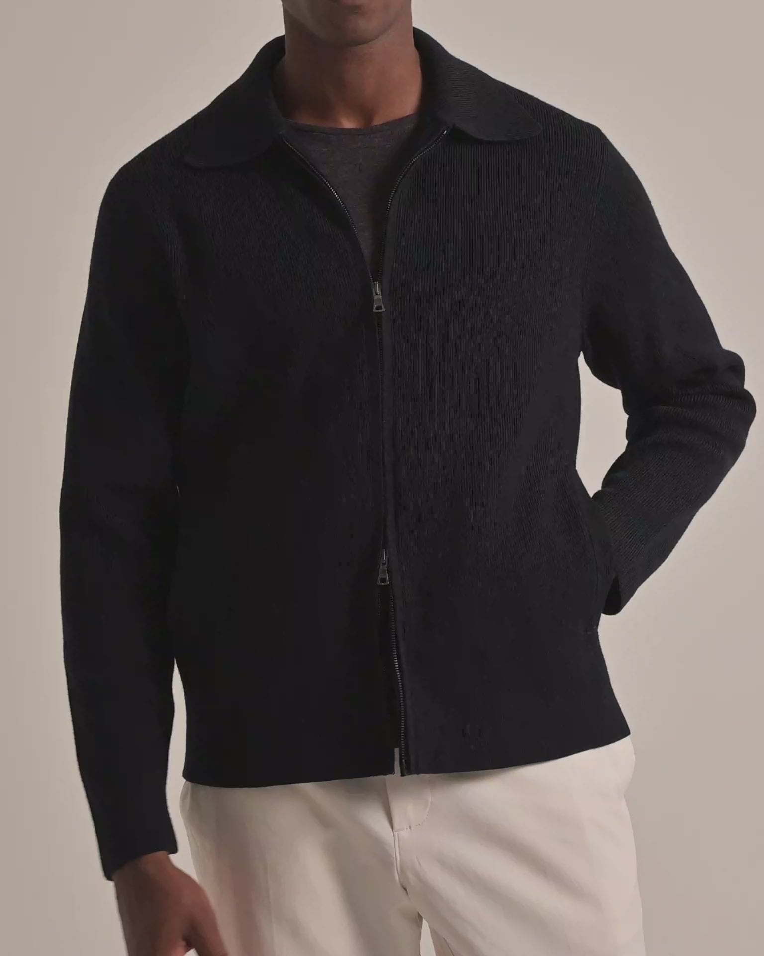 Man wearing black knitted zip-through jacket with dark grey t-shirt underneath and light beige trousers on a plain background