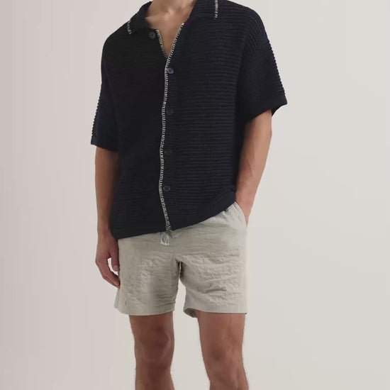 Man wearing crochet stitch shirt in k navy with white detailing, with beige linen shorts