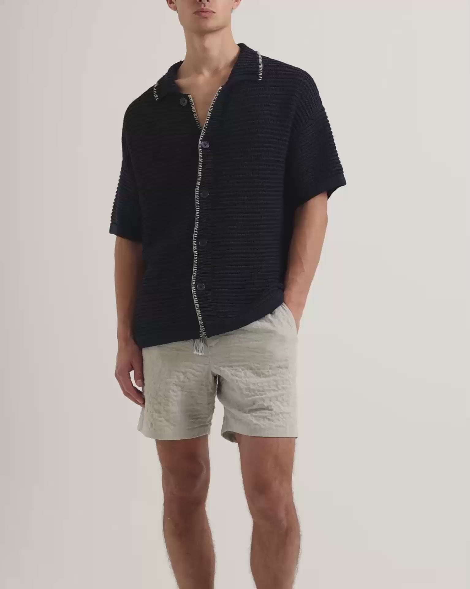 Man wearing crochet stitch shirt in k navy with white detailing, with beige linen shorts