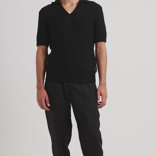 Man wearing black knitted polo shirt with black trousers on a plain background