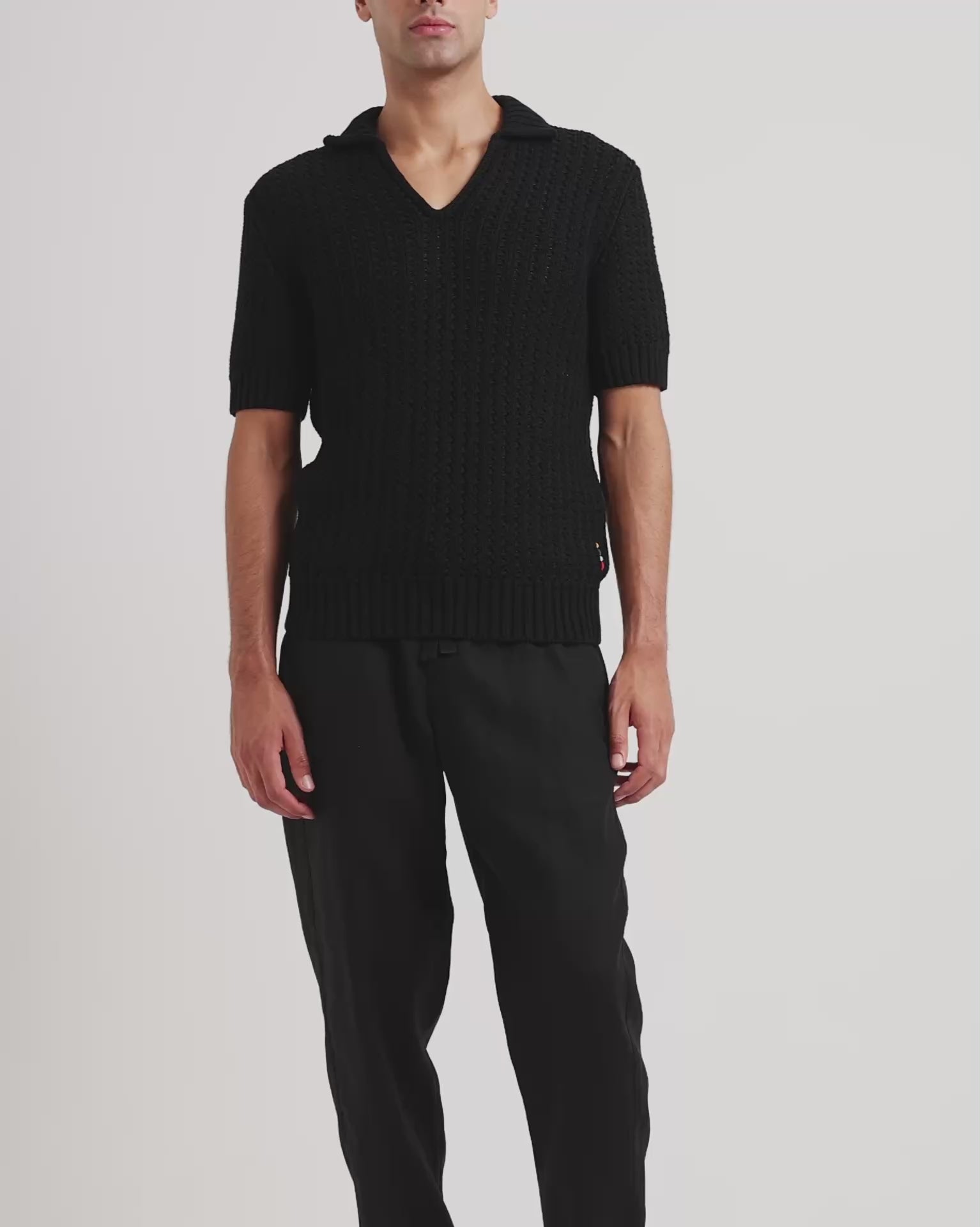 Man wearing black knitted polo shirt with black trousers on a plain background