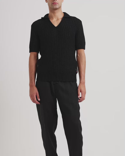 Man wearing black knitted polo shirt with black trousers on a plain background