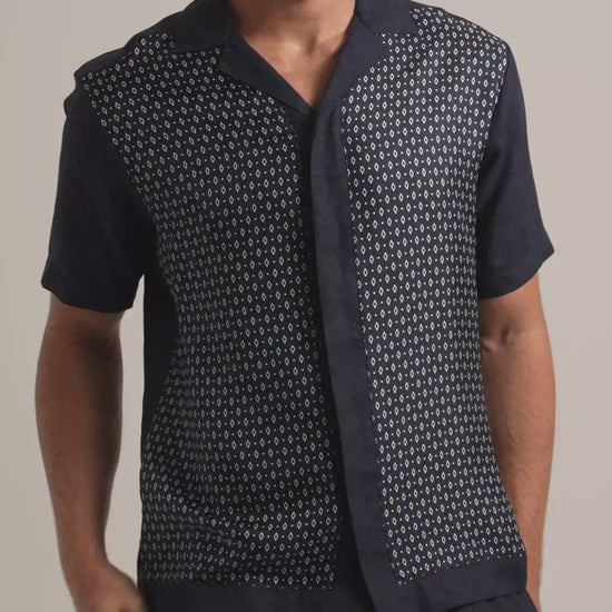 Man wearing silk geometric printed shirt in black and white with dark trousers against a plain background