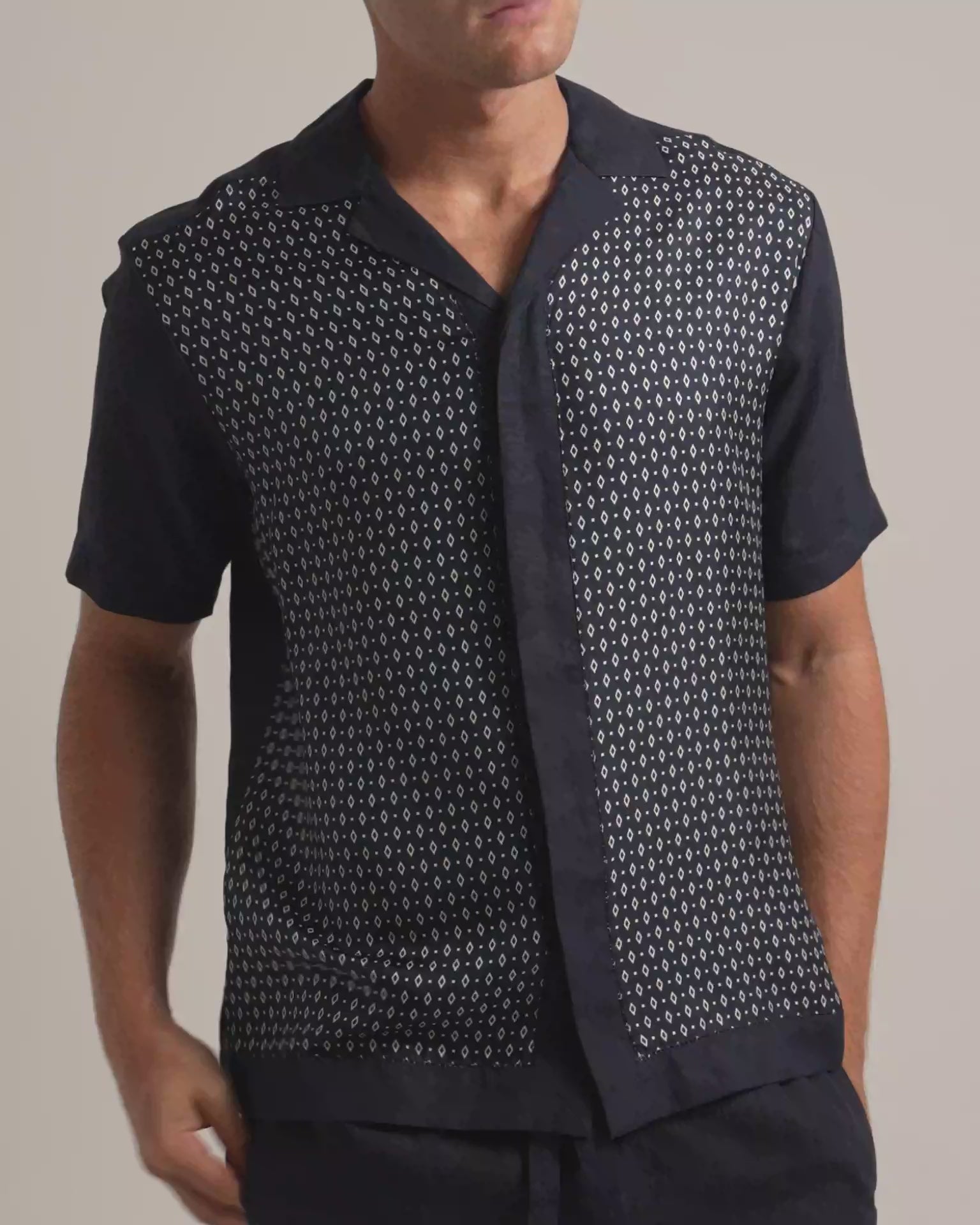 Man wearing silk geometric printed shirt in black and white with dark trousers against a plain background