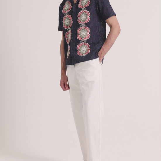 Man wearing navy broderie shirt with white linen trousers on a plain background