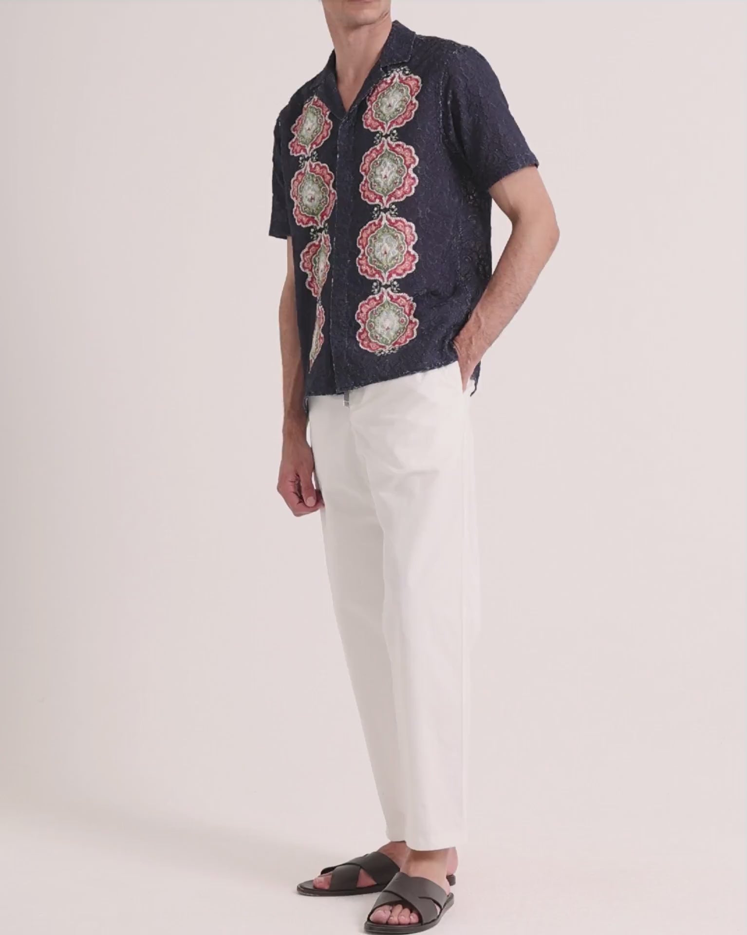 Man wearing navy broderie shirt with white linen trousers on a plain background