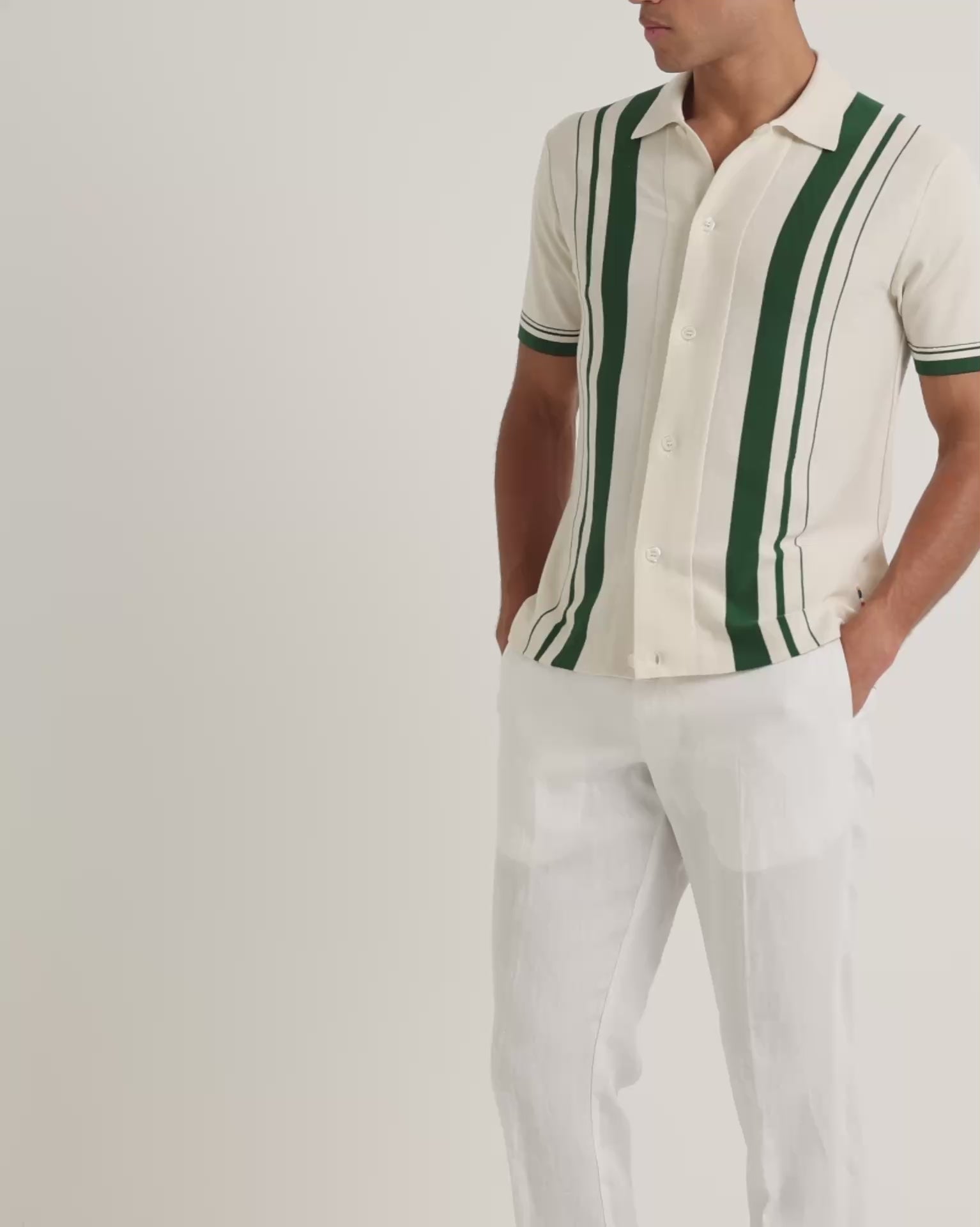 Man wearing light beige shirt with green stripe detail with white trousers on a plain background