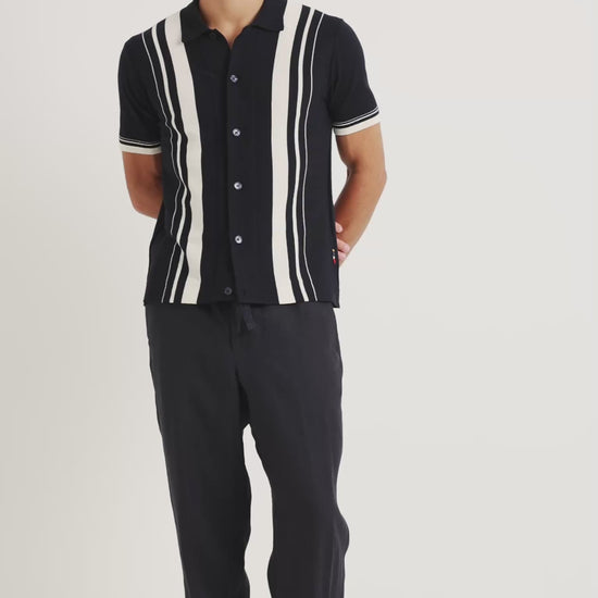 Man wearing black knitted shirt with white stripe detailing with black trousers on a plain background