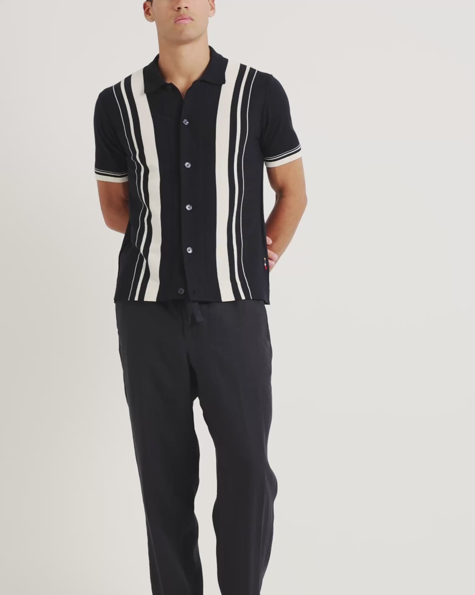Man wearing black knitted shirt with white stripe detailing with black trousers on a plain background