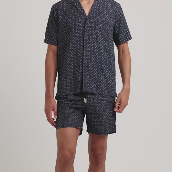 Man wearing navy geometric print shirt with matching swim shorts on a plain background