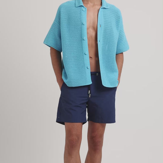 Man wearing blue knitted shirt with navy drawstring swim shorts on a plain background