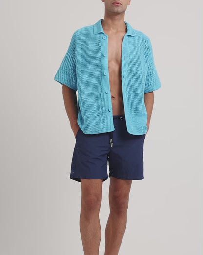 Man wearing blue knitted shirt with navy drawstring swim shorts on a plain background