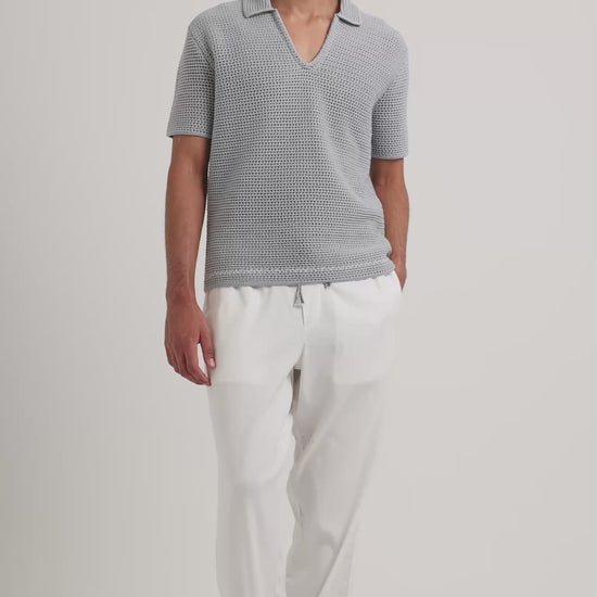 Man wearing light blue crochet polo shirt with white trousers on a plain background
