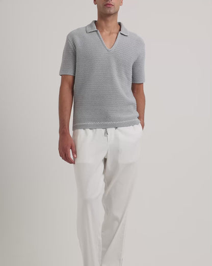 Man wearing light blue crochet polo shirt with white trousers on a plain background