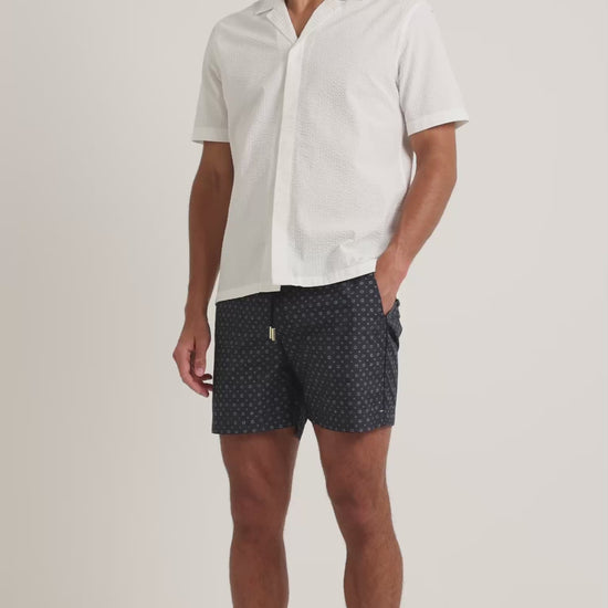 Man wearing white short sleeve shirt with navy geometric print swim shorts on a plain background