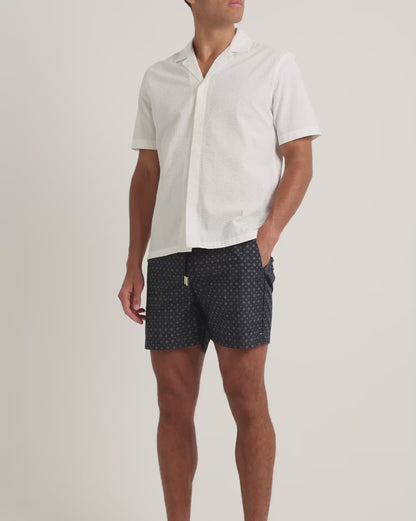 Man wearing white short sleeve shirt with navy geometric print swim shorts on a plain background