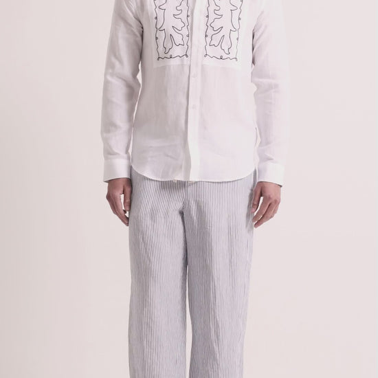 Man wearing linen embroidered shirt with striped trousers on a plain background