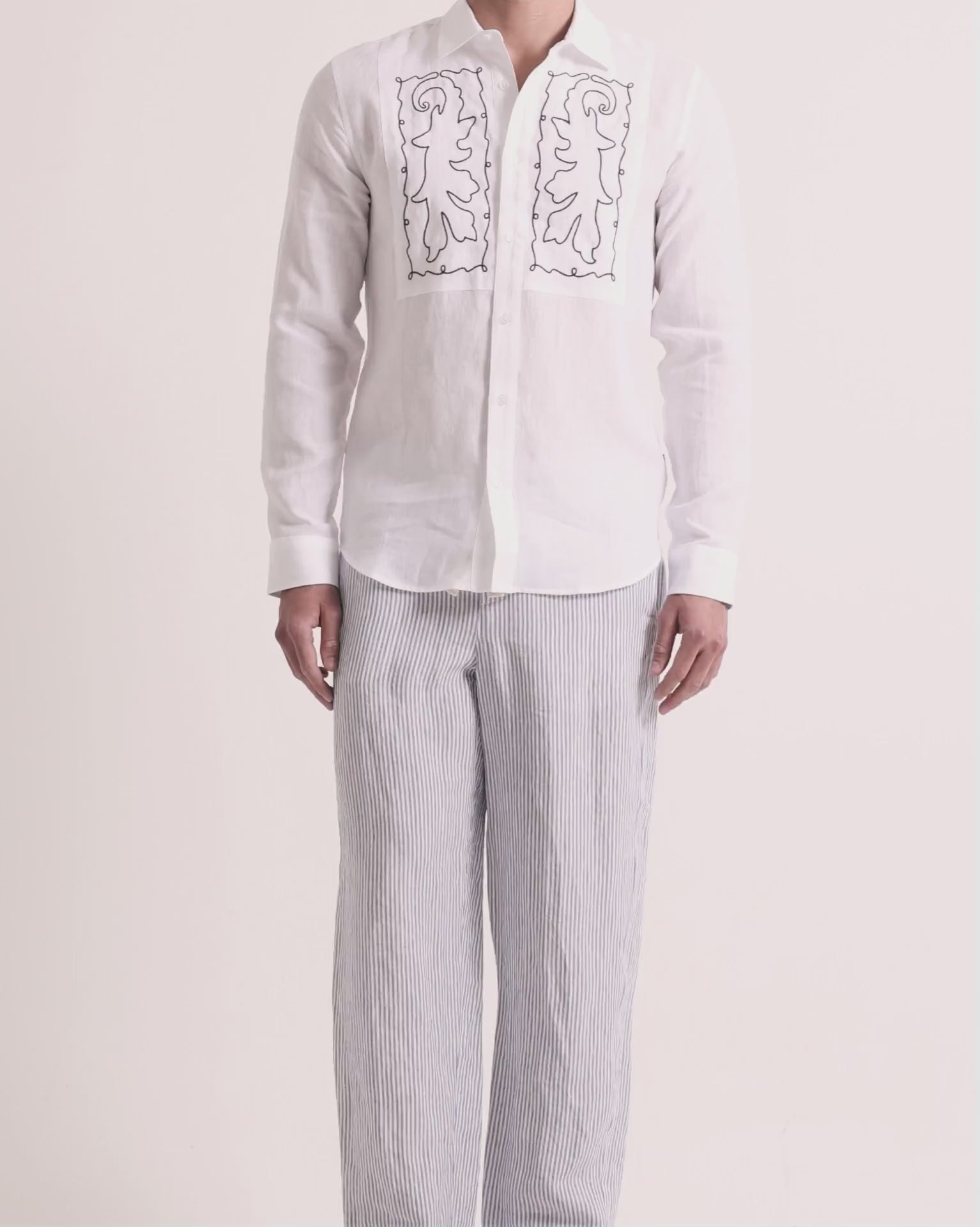 Man wearing linen embroidered shirt with striped trousers on a plain background